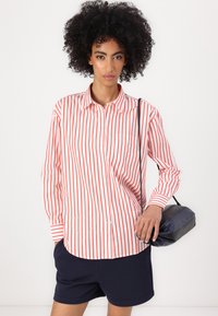Red and white striped button-up shirt with a pocket, worn with navy shorts. Model holds a small navy handbag, showcasing casual attire.