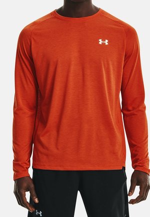 Hombre con camiseta deportiva de manga larga naranja con logo blanco de Under Armour y pantalones cortos negros con logo de Under Armour, de pie con los brazos relajados.