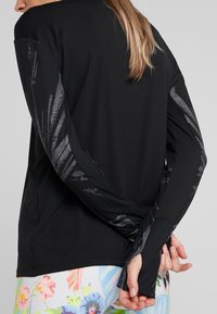 Kobieta w czarnej bluzce z d�ługim rękawem w szare abstrakcyjne wzory na rękawach oraz wielokolorowych legginsach w kwiaty, z rękami złożonymi za plecami.