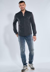 Herren-Hemden in Dunkelgrau mit langen Ärmeln, schlanker Passform, Kentkragen und dezentem Branding. Kombiniert mit hellblauen Jeans und Sneakern.