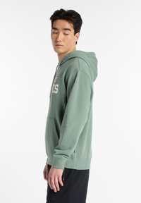 Green cotton hoodie with a front pocket and drawstring hood, featuring white "Athletics" text on the chest, worn over black shorts.