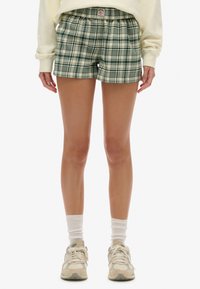 Pantalones cortos de cuadros verdes y crema con cintura elástica, que cuentan con bolsillos laterales y un ajuste holgado. Combinados con calcetines blancos hasta el tobillo y zapatillas deportivas.