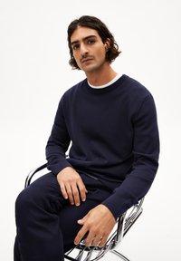 Marineblauwe crewneck trui van zacht materiaal, met ribgebreide manchetten. Model zit op een chroomstoel met ontspannen handplaatsing.