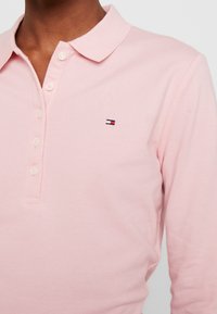 Jasnoróżowa koszulka polo z długim rękawem i kołnierzykiem, z trzema guzikami z przodu oraz małym haftowanym logo na piersi.