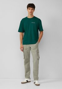 Groene katoenen t-shirt met korte mouwen, ronde hals en witte tekst. Gecombineerd met licht kaki cargobroek en witte sneakers met donkere accenten.