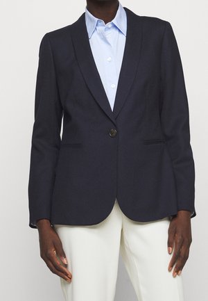 Personne portant un blazer bleu marine foncé avec un seul bouton sur une chemise bleu clair et un pantalon blanc, debout devant un fond uni.