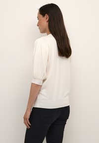 Pull crème à manches courtes bouffantes avec texture côtelée. Ourlet inférieur ajusté, associé à un jean noir. Design simple et épuré.