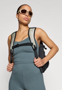 Melnā acu soma uz teala krāsas topa, ar vieglu dizainu un regulējamām siksnām. Modelis nēsā bēšas saulesbrilles.