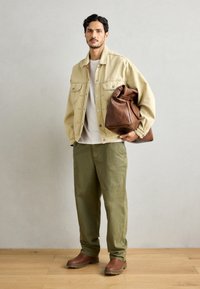 Beige denim jacket, white shirt, olive green pants, brown leather boots, and brown leather bag. Model stands on light wooden floor.