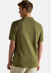 Olivgrünes Poloshirt mit kurzen Ärmeln, Kragen, seitlichen Schlitzen und einer weichen Textur. Einfaches Design, keine sichtbaren Logos oder Muster.