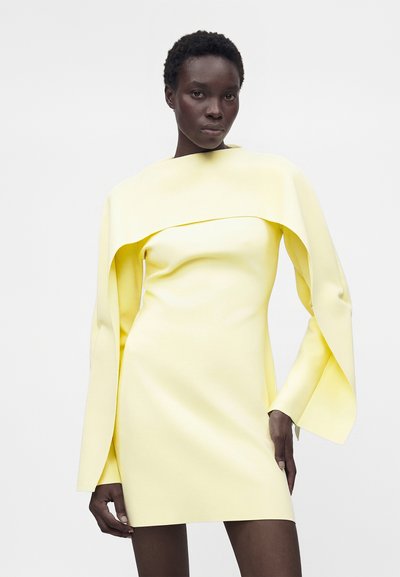 Γυναίκα που φοράει στενή ανοιχτό κίτρινη φορεσιά με δραματικά μακριά μανίκια σαν κάπα, στέκεται μπροστά σε απλό ανοιχτό φόντο.