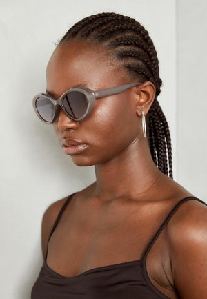 Pilki per dideli akiniai suapvalinta forma ir tamsiais lęšiais. Išskirtinės plastikinės rėmo formos glotnus paviršius. Modelis turi nertas plaukus.