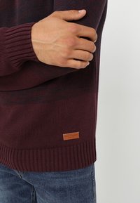 Burgundowy sweter z dzianiny z prążkowanymi mankietami i dołem. Posiada małą, jasnobrązową skórzaną metkę na dolnej stronie. Klasyczny krój i teksturowana tkanina.