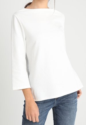 Frau trägt ein strukturiertes weißes Langarmshirt mit einem Bootsausschnitt und blauer Jeans, steht vor einem schlichten hellen Hintergrund.