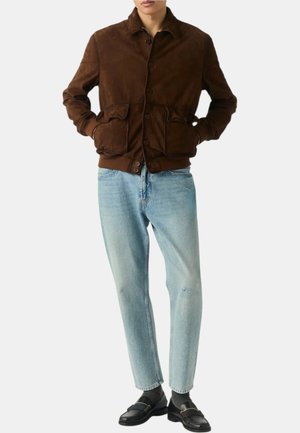 Giacca in suede marrone con due tasche anteriori, chiusura con bottoni e colletto. Indossata sopra jeans azzurri chiari e scarpe nere senza lacci.