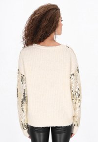 Crèmekleurige trui met een gebreid lichaam met textuur en mouwen met metalen paillettenaccenten. Heeft een losse pasvorm en een ronde hals.
