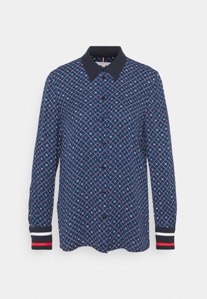 Chemise bleu marine à manches longues avec col noir, boutonnage devant, motif géométrique en treillis avec lettres, et poignets rayés en noir, blanc et rouge.