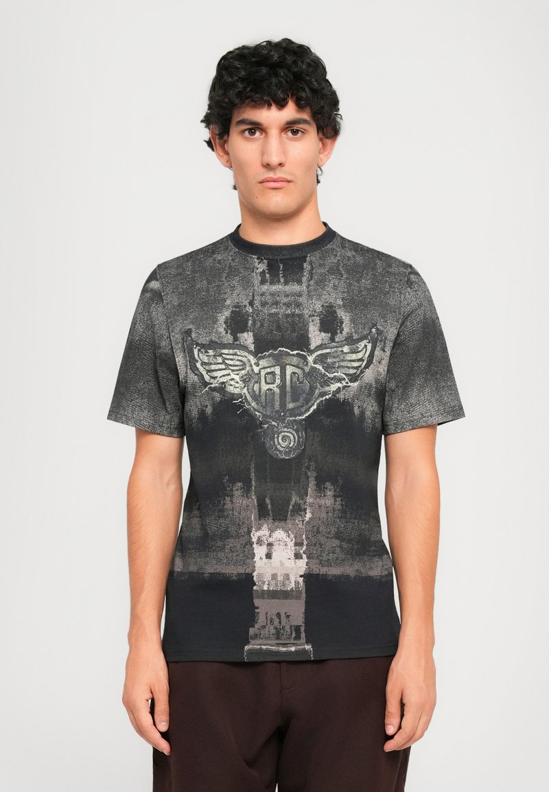 Young man with curly dark hair wears a black and gray abstract patterned t-shirt with a winged emblem on the chest, standing against a plain backdrop.