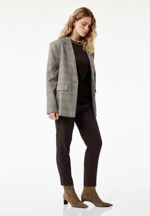 Kvinde med bølget blondt hår iført en grå ternet blazer, sort top og bukser samt brune ruskindsstøvler, stående mod en hvid baggrund.