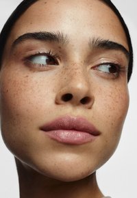 Närbild av ett ansikte med ljusbrun hy, framträdande fräknar, fylliga rosa läppar, definierade ögonbryn och subtilt ögonsmink mot en ljus bakgrund.
