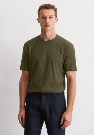 Olivegrünes kurzärmliges T-Shirt aus weichem, strukturiertem Stoff mit Rundhalsausschnitt. Kombiniert mit dunkelblauen, plissierten Hosen.