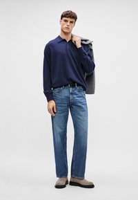 Ναυτικό πουλόβερ πόλο, άνετη εφαρμογή, μακριά μανίκια. Συνδυασμένο με ανοιχτό μπλε τζιν καμπάνα και μπεζ κοτσίδα. Γκρι μπουφάν ριγμένο στον ώμο.