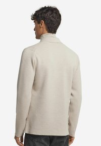 Ein Mann, der von hinten zu sehen ist, trägt einen beigefarbenen Pullover mit langem Arm und hohem Kragen sowie eine dunkle Jeans vor einem schlichten weißen Hintergrund.