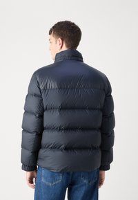 Giacca puffer navy con design trapuntato, colletto alto e polsini elasticizzati. Il materiale è lucido, liscio e spesso, offrendo calore e isolamento.