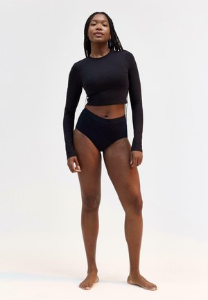 Donna in piedi a piedi nudi su sfondo semplice, che indossa un crop top nero a maniche lunghe e biancheria intima nera a vita alta.
