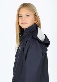 Giacca impermeabile blu navy con collo alto, dettagli grigi con cappuccio, bottoni a pressione e logo sul petto. Tessuto liscio e leggero.