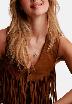Lächelnde Frau mit langen blonden Haaren, die eine braune Wildlederweste mit Fransen und eine silberne Halskette mit einem grünen Froschanhänger trägt.