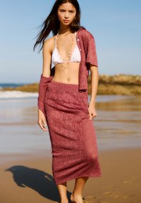 Bordeaux gebreide maxi-rok met een ruitpatroon, gecombineerd met een witte driehoek bikini top en een doorschijnende bijpassende omslagdoek. Strandsetting.