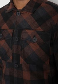 Brązowo-czarna koszulowa kurtka w kratę z kołnierzem, dwoma kieszeniami na piersi i czarnymi guzikami, wykonana z teksturowanej tkaniny.