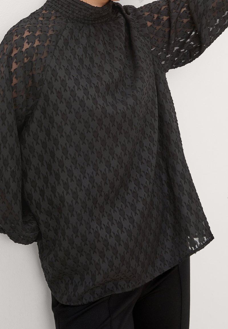 Blouse noire en tissu transparent avec des manches longues bouffantes et un motif pied-de-poule texturé, portée avec un pantalon noir, sur un fond clair uni.