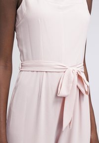 Vaaleanpunainen hihaton haalari, jossa on teksturoitu kangas. Se sisältää muotoillun vartalon yläosan ja sidottavan vyötäröyksityiskohdan, joka antaa kireän siluetin.