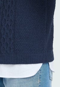 Mørkeblå strikket genser med et teksturert kabelmønster på siden, kombinert med en hvit skjorte under og lyse denim jeans.