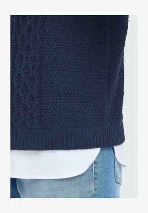 Mørkeblå strikket genser med et teksturert kabelmønster på siden, kombinert med en hvit skjorte under og lyse denim jeans.
