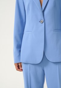 Světle modrý tailored blazer s klopovým límcem, zapínáním na jeden knoflík a dvěma předními kapsami. Hladká textilie, těsně přiléhající střih.