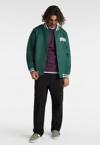 Vihreä pommikurkkutakki, jossa valkoiset raidat kauluksessa ja hihansuissa, hopeiset napit ja teksturoitu graafinen yksityiskohta rinnassa. Mustat cargo-housut.