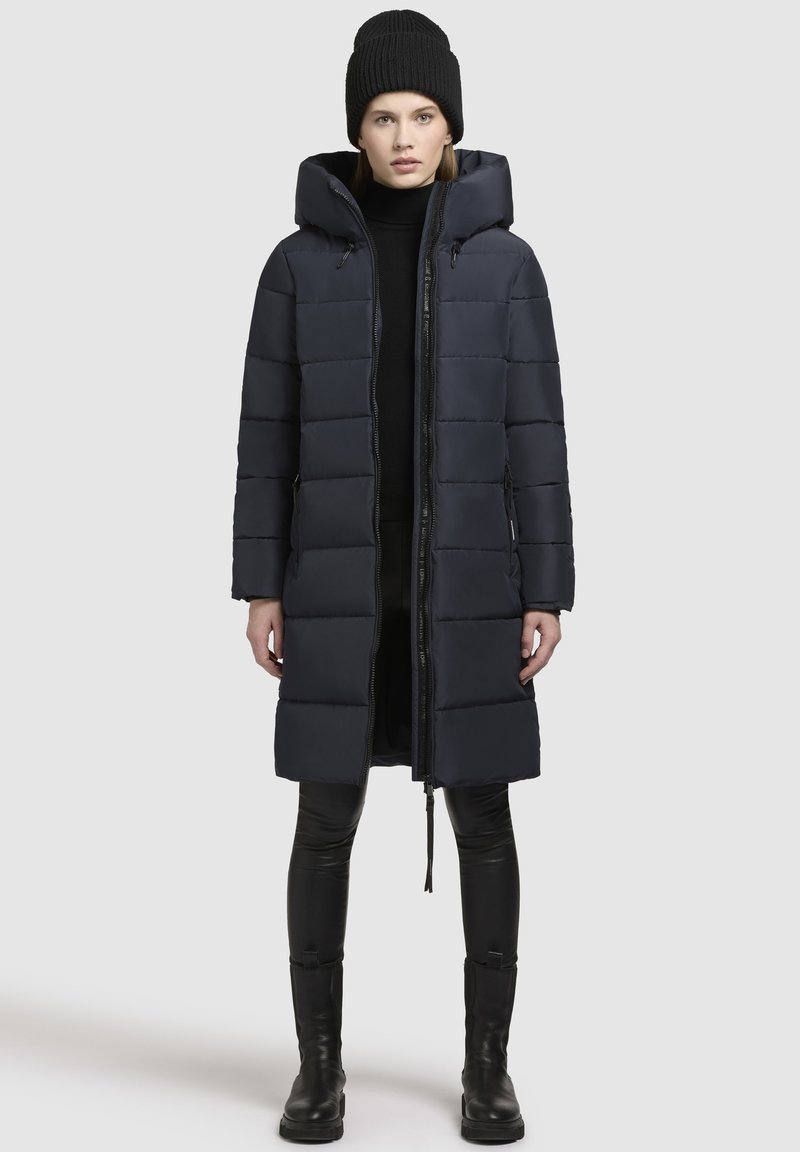 Tengeri kék párnázott hosszú kabát, kapucnival, elől cipzáras záródással és oldalsó zsebekkel. Fekete leggingsszel és térd magas csizmával párosítva. Kötött fekete sapka.