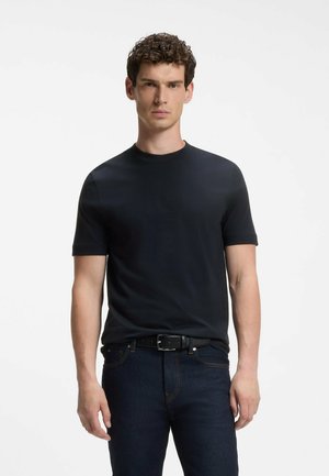 Mann mit lockigem Haar, der ein schlichtes schwarzes kurzärmeliges Shirt, dunkle Jeans und einen schwarzen Gürtel trägt, steht vor einem einfarbigen weißen Hintergrund.