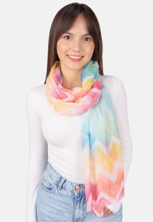 Young woman smiling, wearing a white long-sleeve top, light blue jeans, and a colorful zigzag patterned scarf wrapped around her neck.