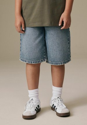 Denimshorts med løs pasform og frynset kant, kombineret med hvide crew-sokker og hvide/sølvfarvede sports sko med sorte detaljer.