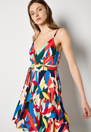 Kobieta ubrana w kolorową, abstrakcyjną sukienkę bez rękawów z cienkimi ramiączkami i kopertowym dekoltem w kształcie litery V.