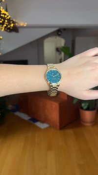 Montre-bracelet en métal doré avec un cadran rond turquoise, des marqueurs d'heure argentés et un bracelet à maillons. Affiche l'heure avec deux aiguilles rouges.