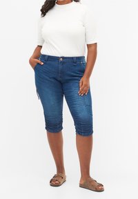 Capris de mezclilla azul oscuro con un diseño ceñido, que presentan bolsillos laterales y un dobladillo fruncido. Combinados con una blusa blanca de manga corta y sandalias.