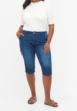Capris de mezclilla azul oscuro con un diseño ceñido, que presentan bolsillos laterales y un dobladillo fruncido. Combinados con una blusa blanca de manga corta y sandalias.