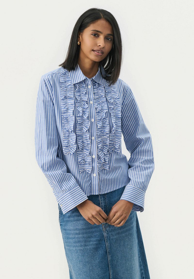 Blå och vit randig skjorta med volangdetalj framtill, knapplist och långa ärmar. Bärs med en jeanskjol. Slät tygtextur.