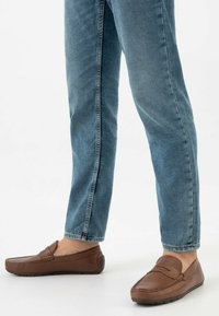 Mocassini in pelle marrone con design a mocassino, caratterizzati da dettagli cuciti e suole in gomma, abbinati a jeans di denim azzurro chiaro.