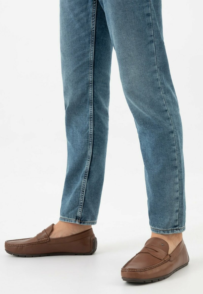 Brązowe skórzane loafersy w stylu mokasynów, z detalami przeszycia i gumowymi podeszwami, połączone z jasnoniebieskimi dżinsami.